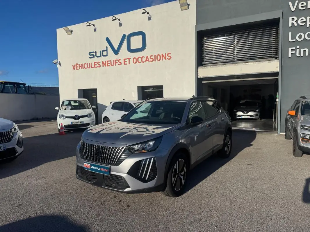 Peugeot 2008 Hybrid gris en 3/4 avant droit avec barres de toit et jantes alliage 17 pouces, stationné devant un garage.