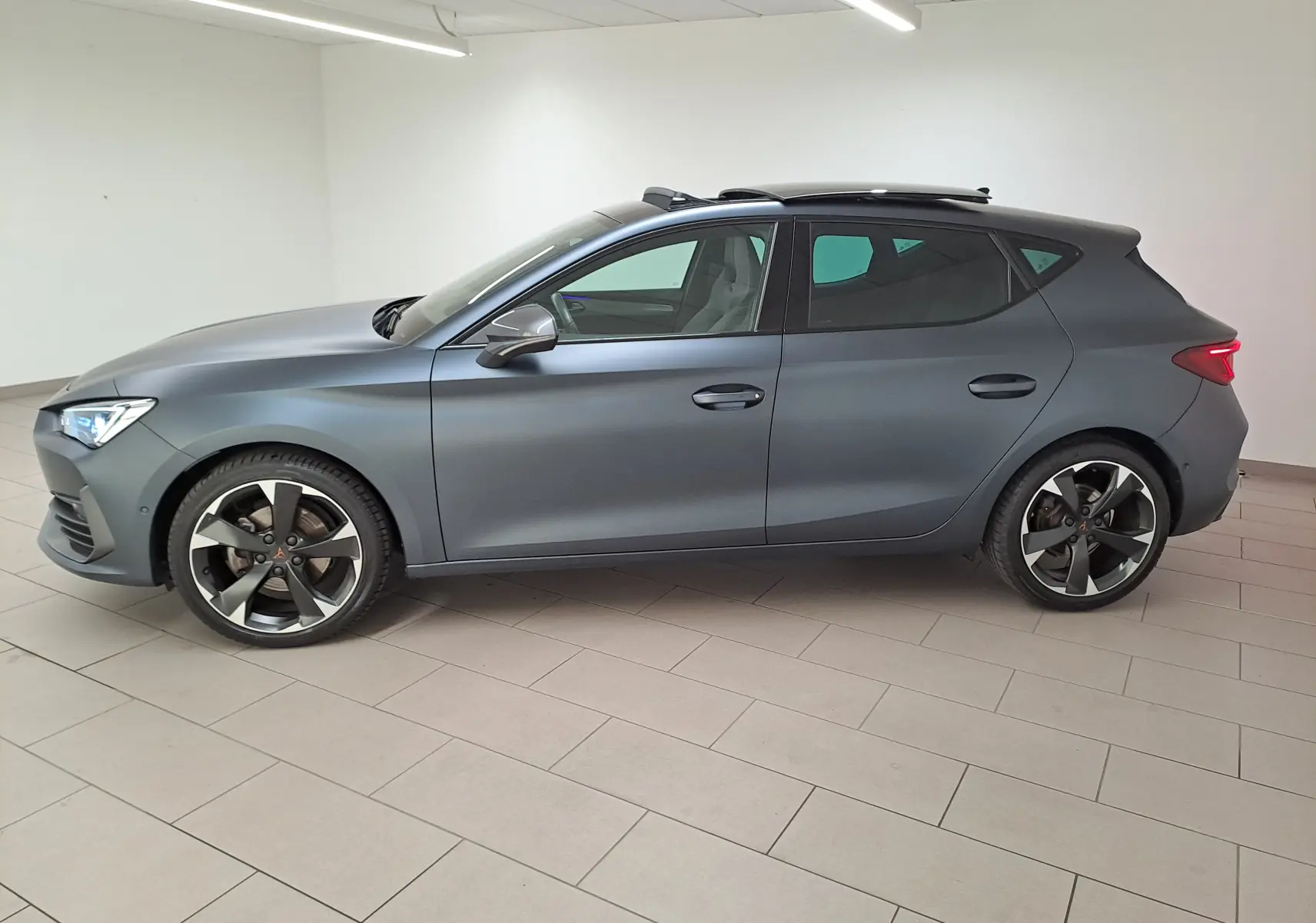 Vue de profil côté gauche d'une CUPRA Leon 2024 gris magnétique mat avec toit ouvrant panoramique en intérieur showroom.