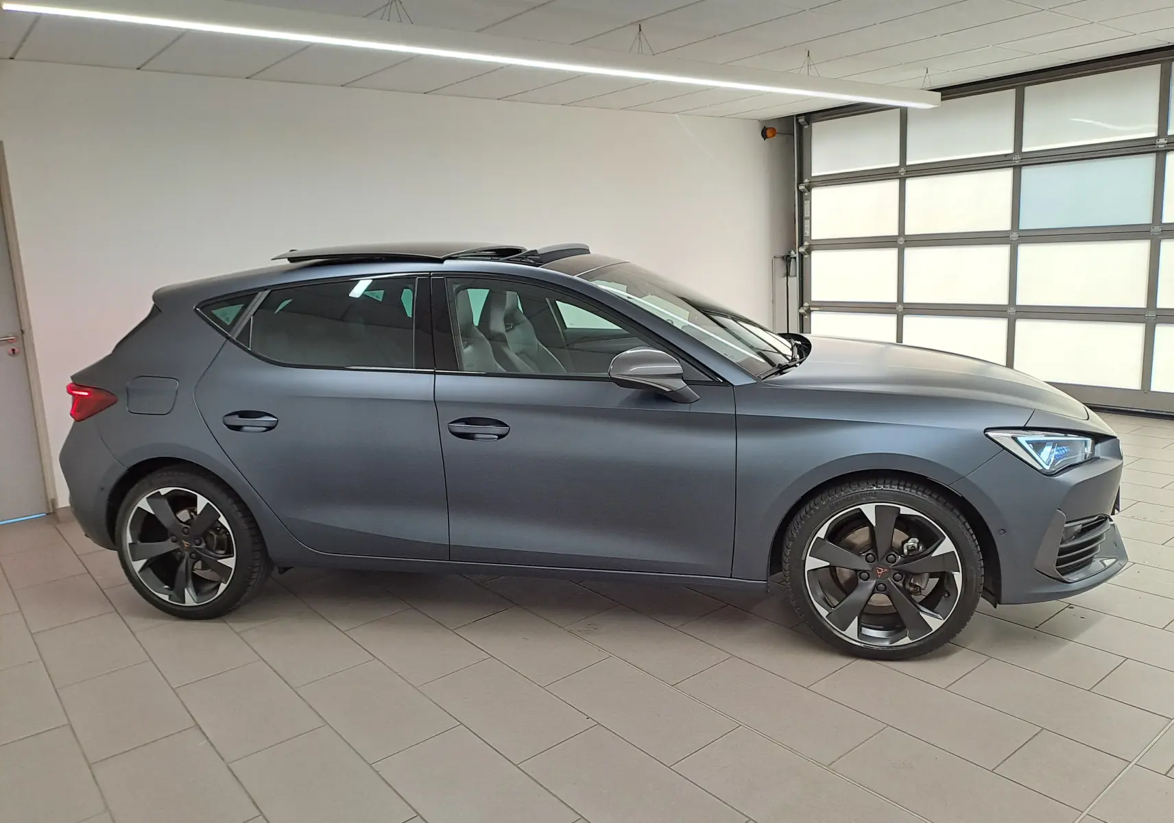 Vue de profil côté gauche d'une CUPRA Leon 2024 gris magnétique mat avec toit ouvrant panoramique en intérieur.