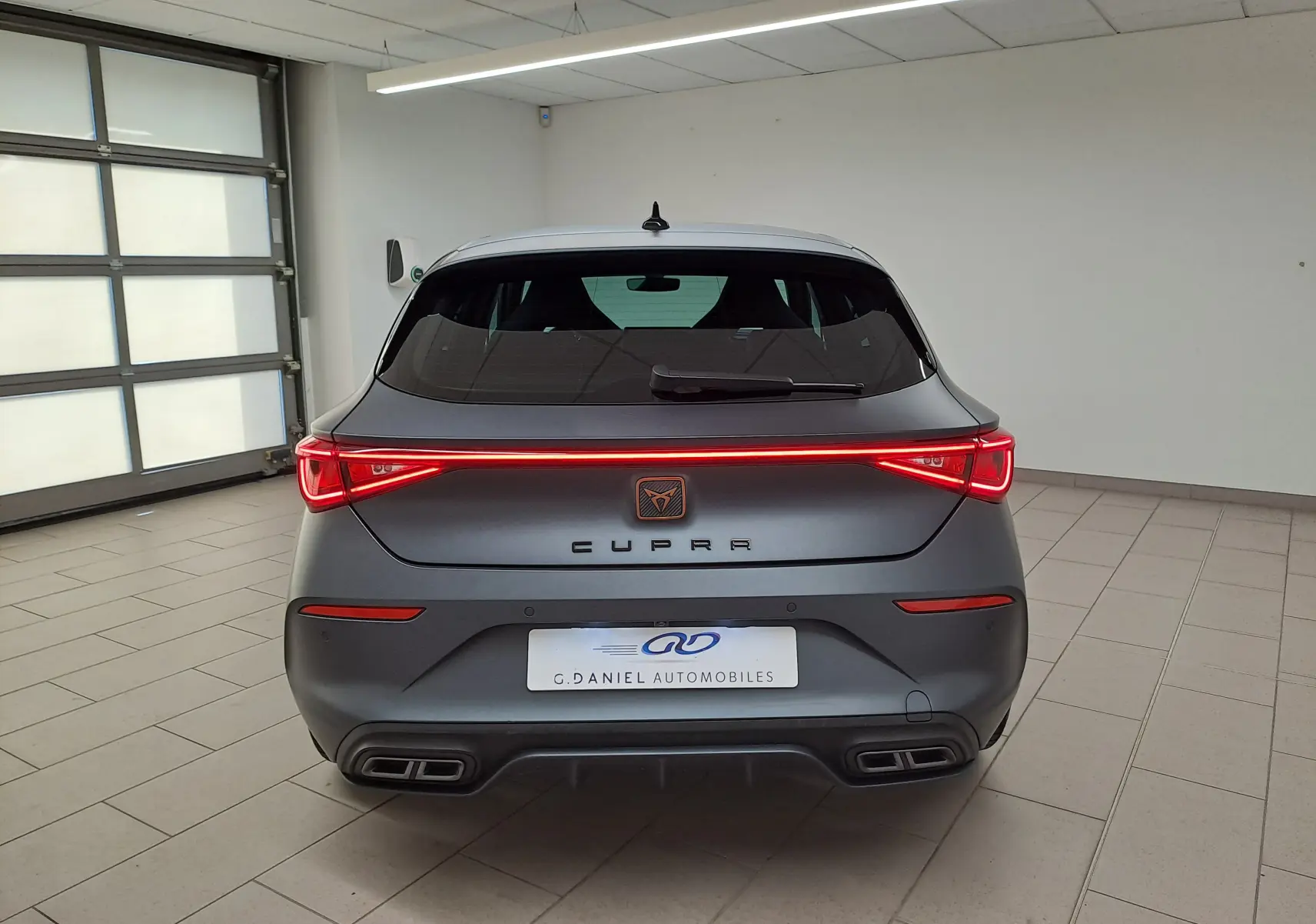 Vue arrière d'une CUPRA Leon 2024 gris magnétique mat avec feux LED allumés et double sortie d'échappement carrée.