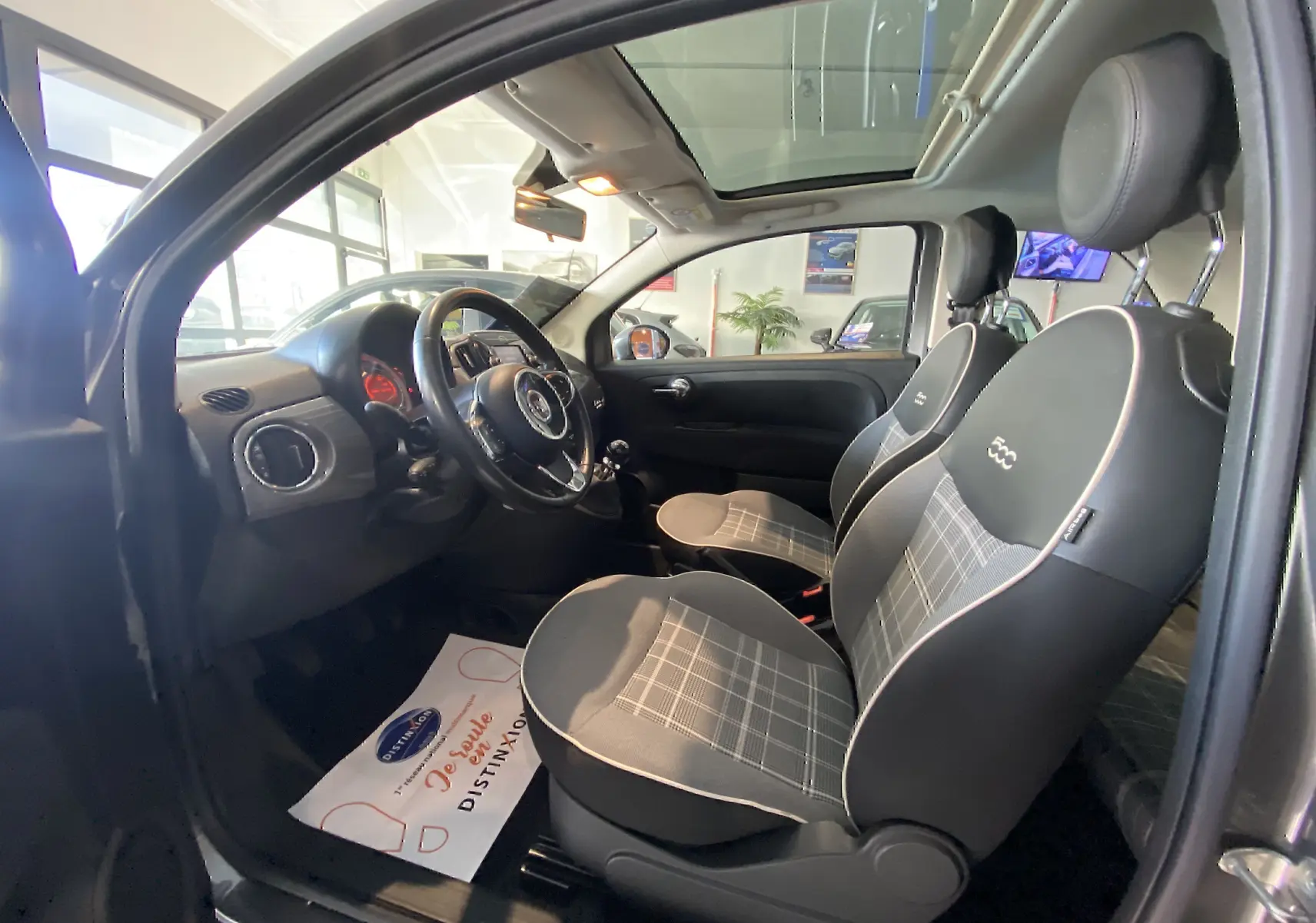 Vue intérieure côté conducteur d'une Fiat 500 Lounge 2019 gris foncé avec sièges noirs à motifs à carreaux et toit ouvrant.