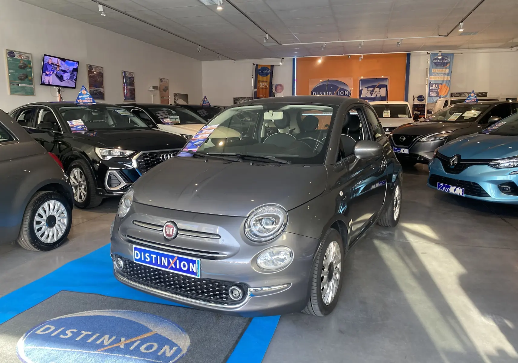 Vue 3/4 avant d'une Fiat 500 gris foncé exposée en showroom avec plaque Distinxion visible.