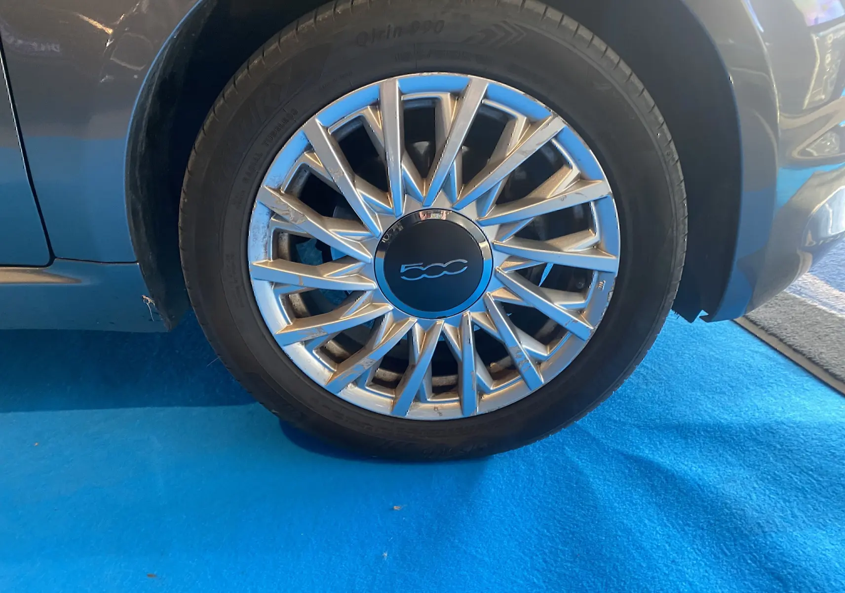 Gros plan sur la roue avant droite d'une FIAT 500 gris foncé avec jante argentée à rayons fins sur sol bleu.