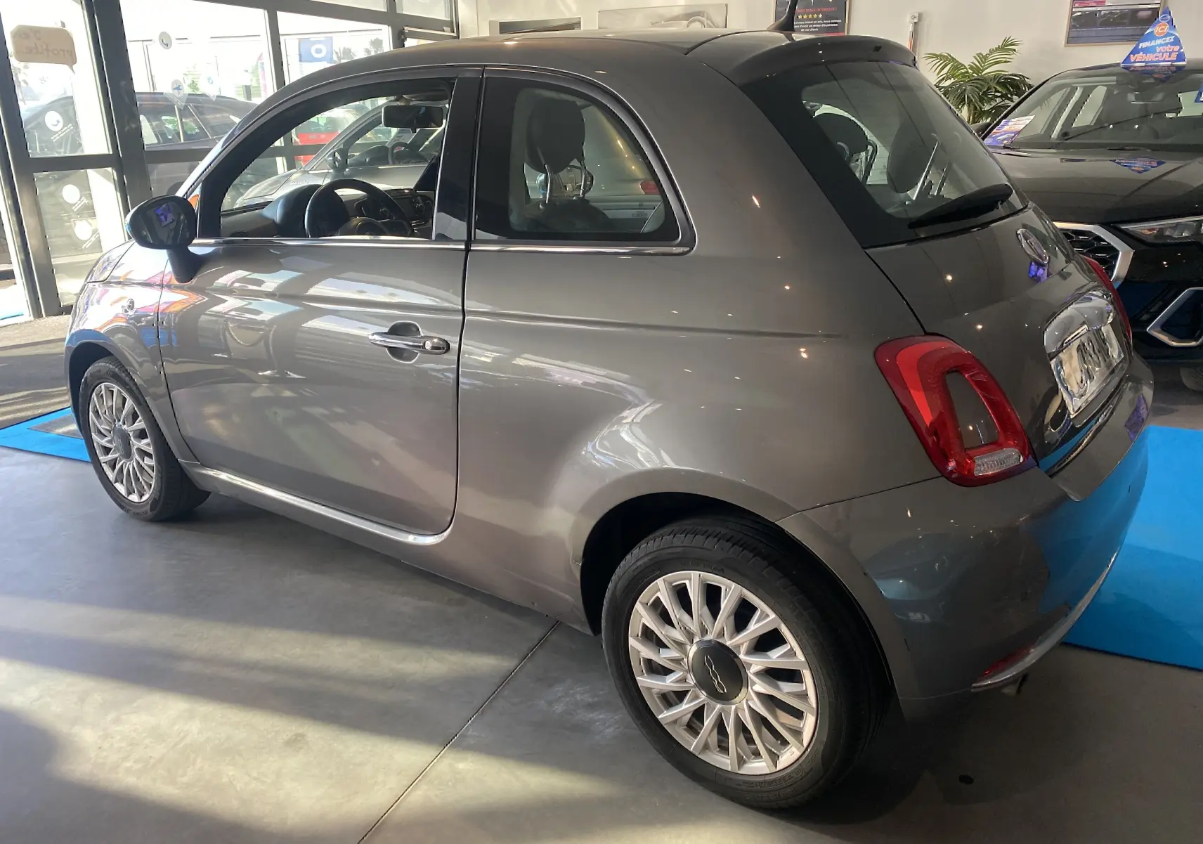 Vue 3/4 arrière droite d'une FIAT 500 Lounge gris foncé avec toit noir, en intérieur showroom lumineux.