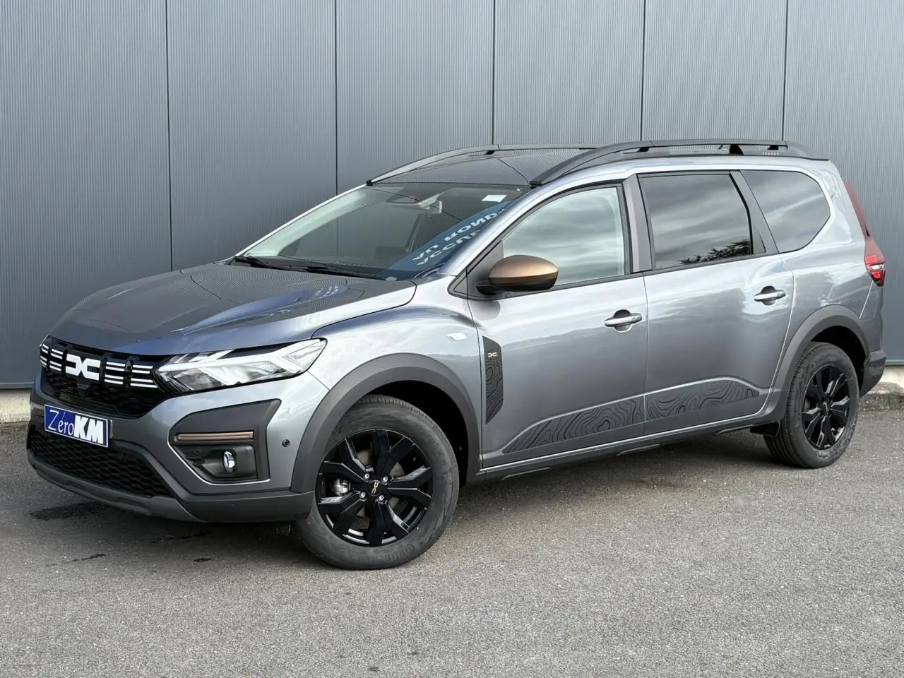 Vue 3/4 avant droit du Dacia Jogger Gris Schiste avec jantes noires et barres de toit modulaires.