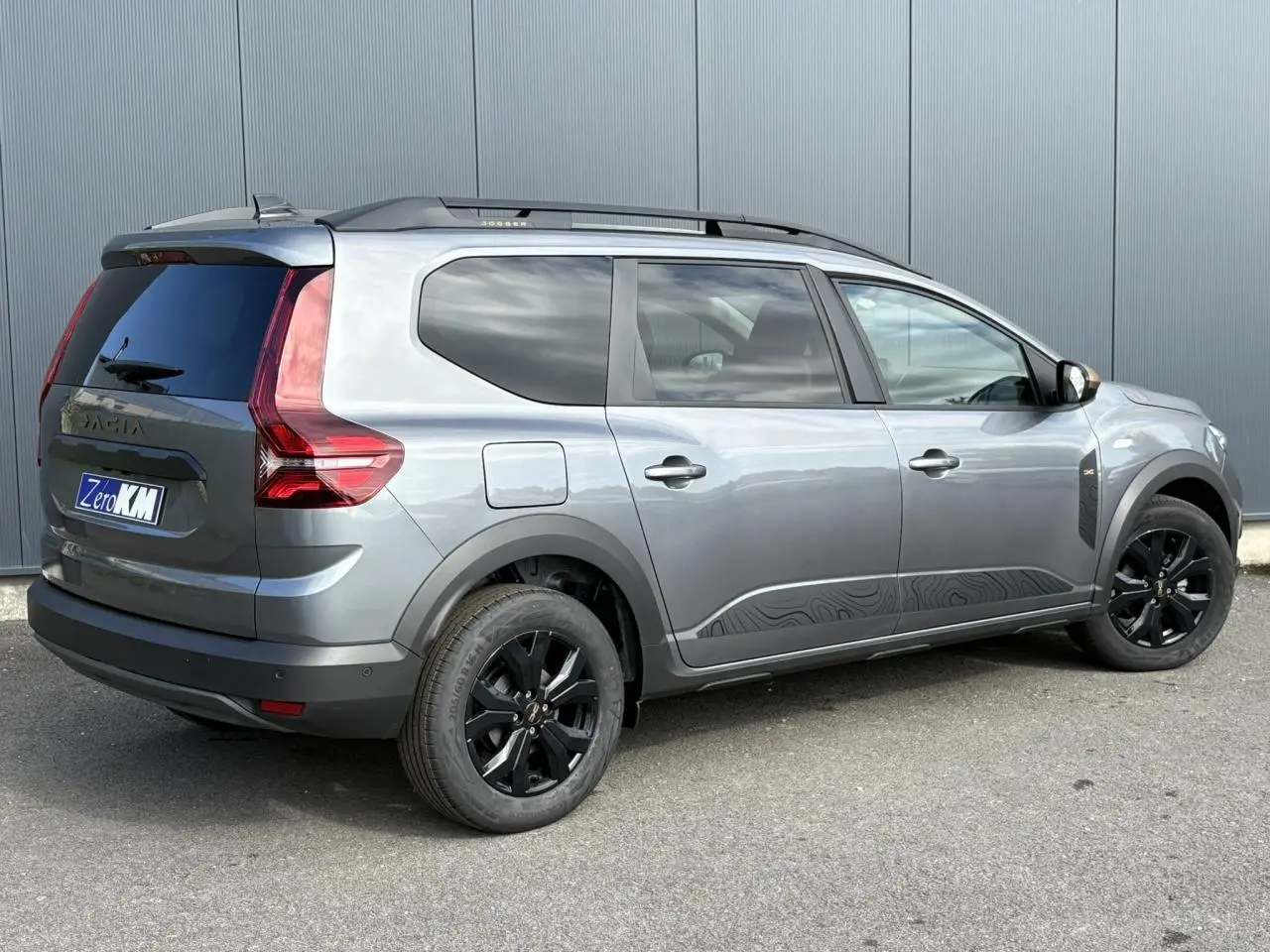 Vue 3/4 arrière droite d'un Dacia Jogger gris schiste avec jantes noires et barres de toit modulaires.