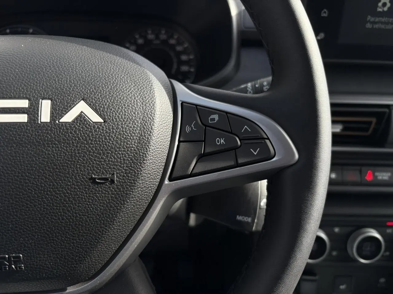 Gros plan sur le volant noir de la Dacia Jogger 2024 avec commandes multifonctions et tableau de bord en arrière-plan.