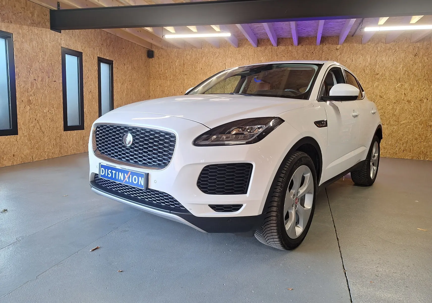 Jaguar E-PACE blanc vue 3/4 avant droit dans un garage avec calandre distinctive et phares LED allumés.