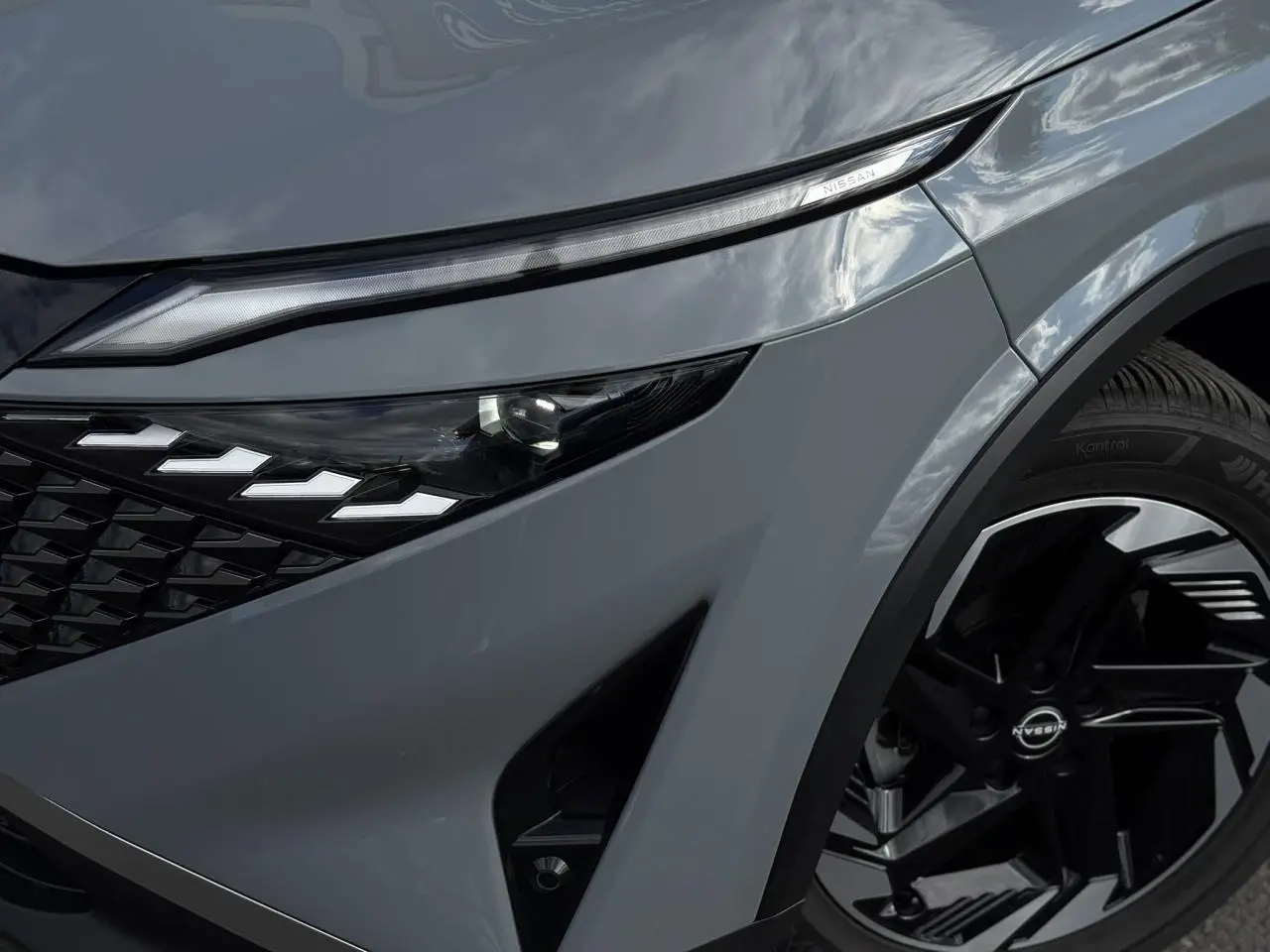 Gros plan sur le phare avant droit et la jante alliage noire du Nissan Qashqai gris Ceramic 2025.