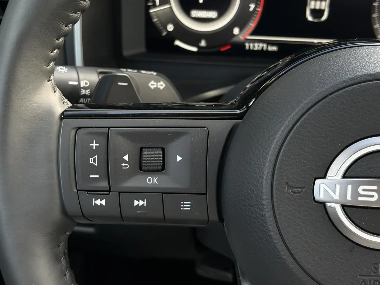 Gros plan sur les commandes gauche du volant cuir du Nissan Qashqai 2025, avec tableau de bord numérique en arrière-plan.