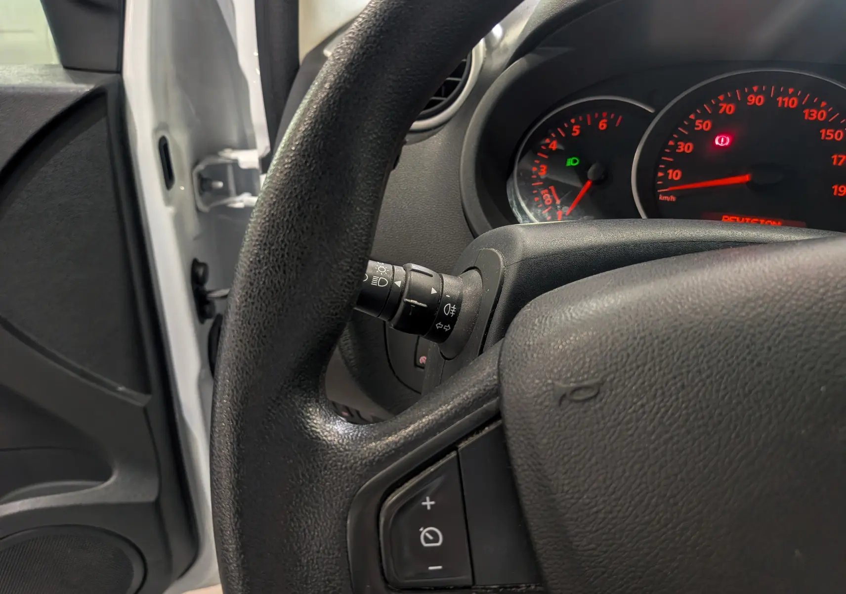 Vue intérieure rapprochée du volant et du tableau de bord du Renault Kangoo Express blanc, avec porte ouverte côté gauche visible.