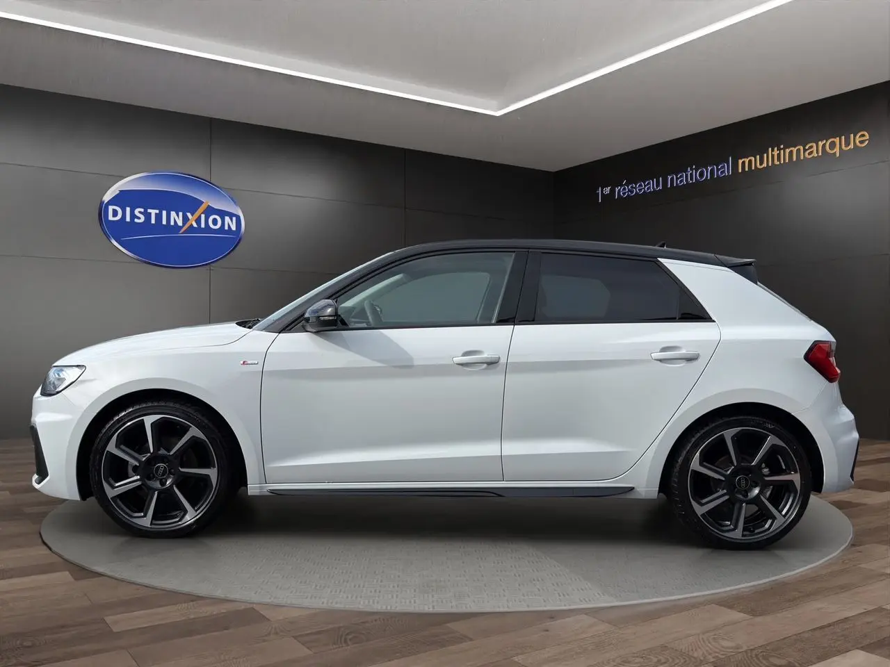 Profil latéral gauche d'une Audi A1 Sportback blanche avec toit noir et jantes alliage distinctives en exposition showroom.