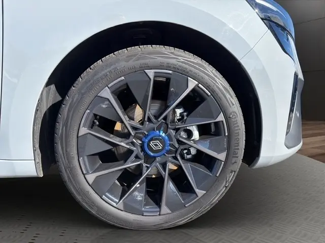 Gros plan sur la roue avant droite d'une Renault Clio gris clair, jante noire au design géométrique avec logo central bleu.