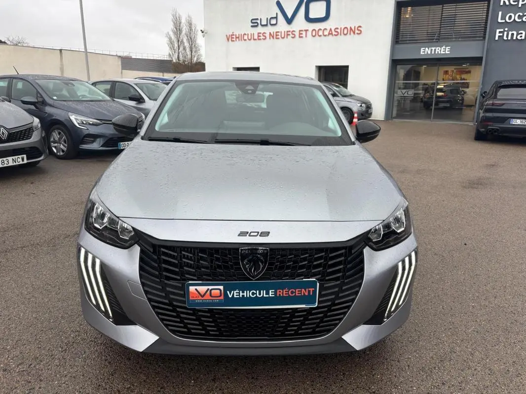 Vue frontale d'une Peugeot 208 gris clair 2025 avec feux diurnes LED en forme de griffes et calandre noire distinctive.