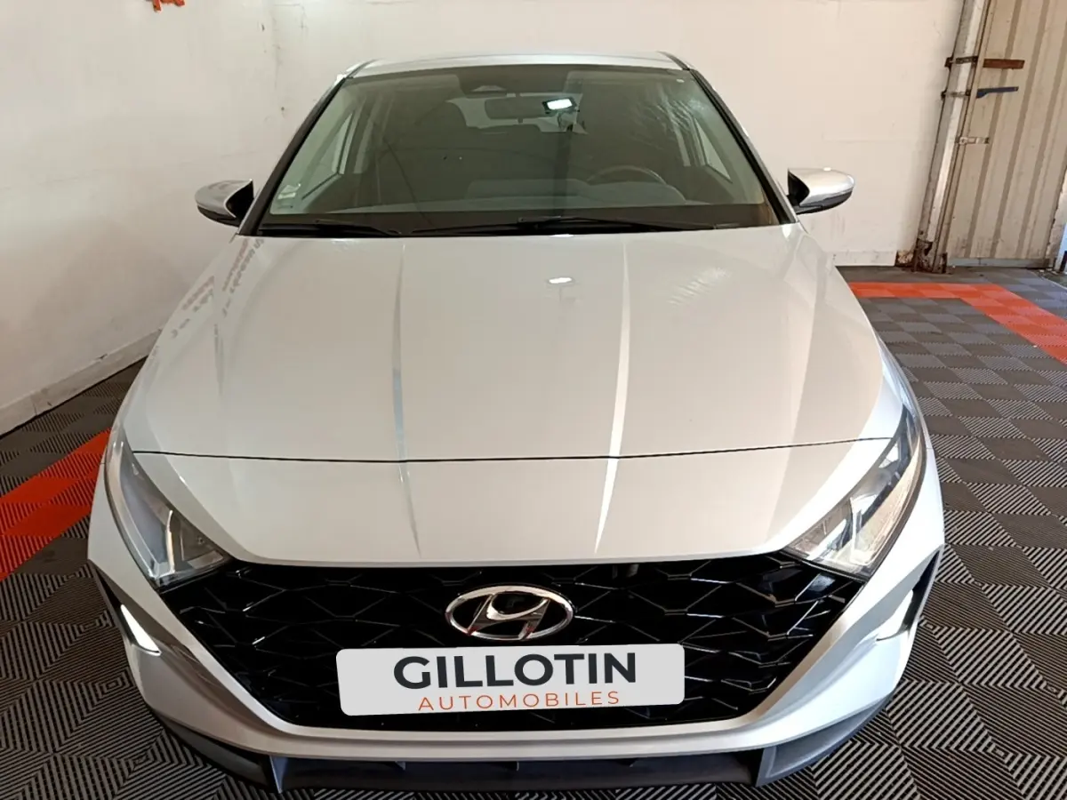 Vue avant d'une Hyundai i20 gris clair 2022 avec calandre noire et phares LED, stationnée en intérieur.