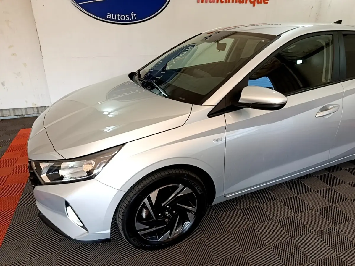 Vue 3/4 avant droit d'une Hyundai i20 gris clair avec jantes noires et badge hybride 48V visible sur l'aile.