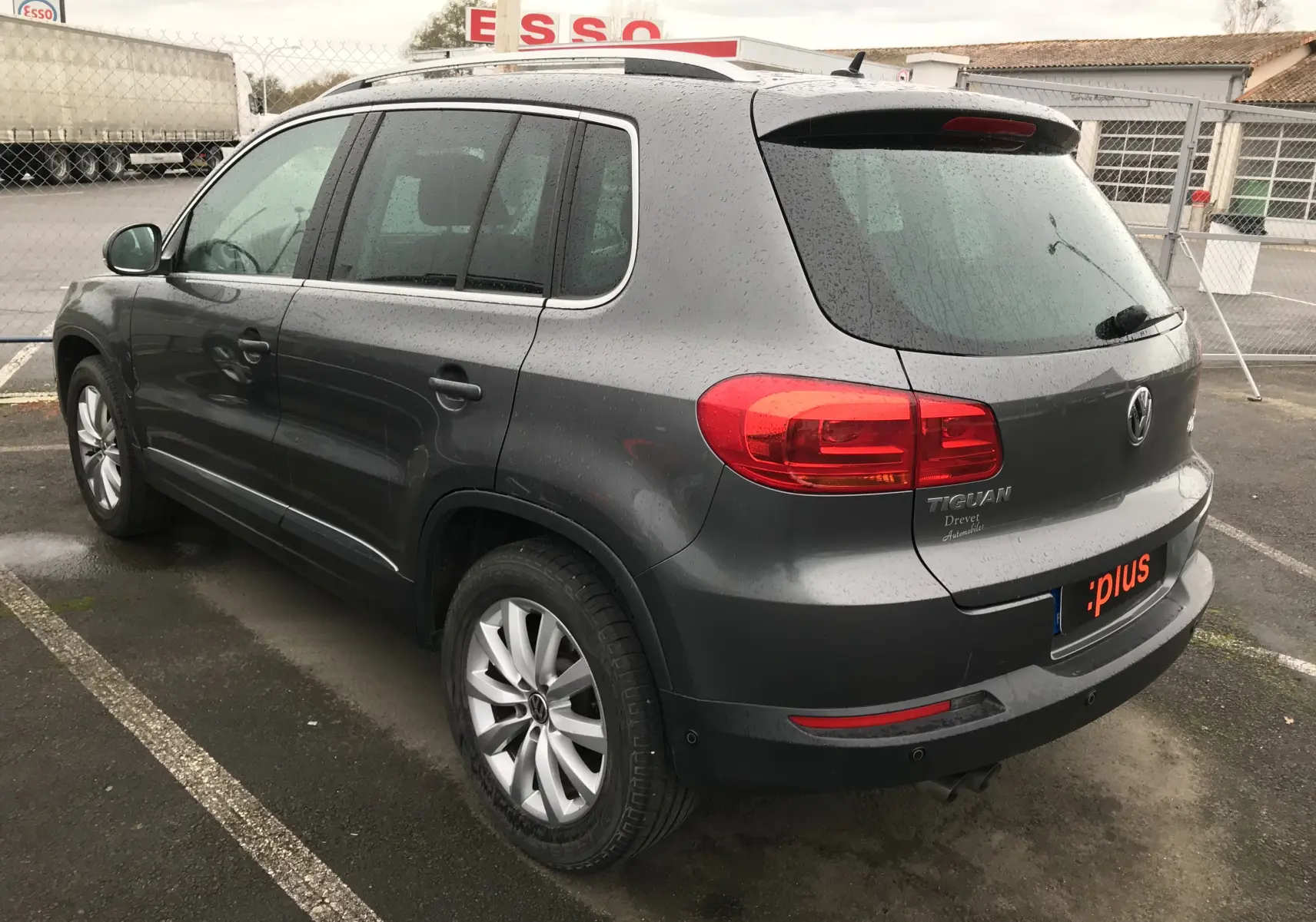 Vue 3/4 arrière gauche d'un Volkswagen Tiguan Sportline gris foncé 2012 avec jantes alliage et feux arrière rouges.