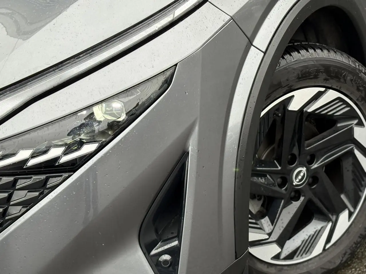 Gros plan sur l'avant droit gris Squale du Nissan Qashqai 2024, mettant en valeur le phare LED et la jante alliage 18 pouces.