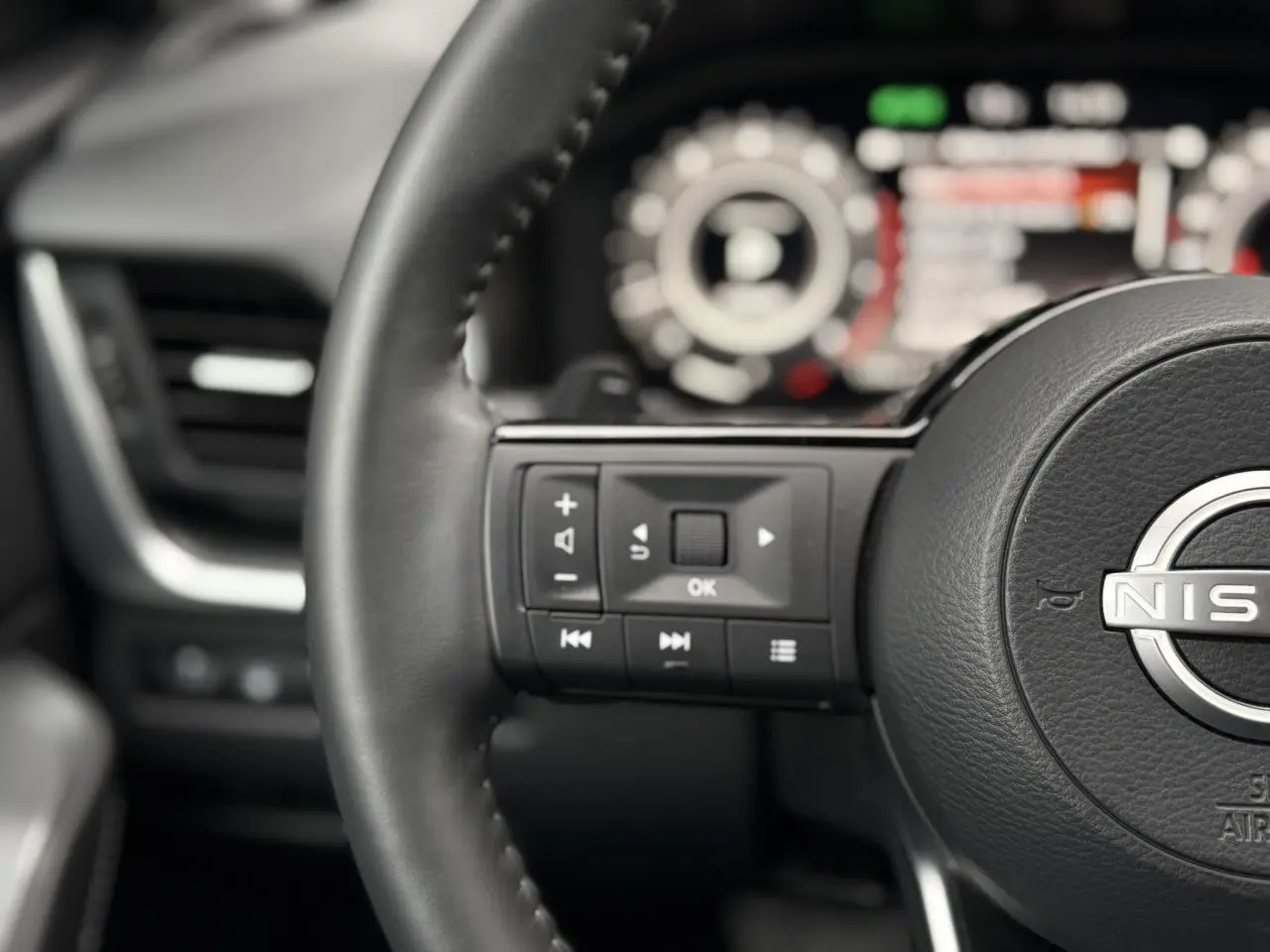 Gros plan sur le volant cuir noir du Nissan Qashqai 2024 avec commandes multifonctions et tableau de bord numérique flou.