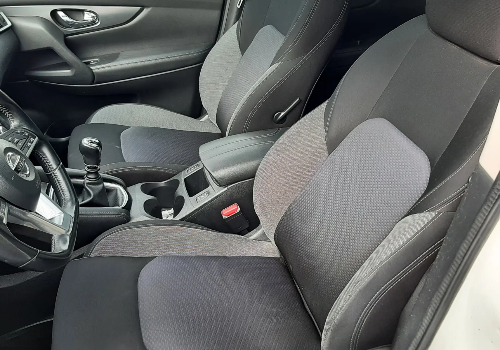 Vue intérieure côté conducteur du Nissan Qashqai blanc 2018, sièges tissu gris et noir avec levier de vitesse manuel visible.