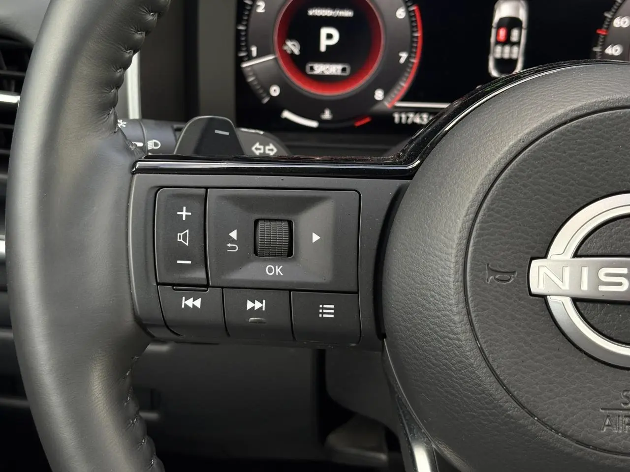 Gros plan sur les commandes gauche du volant cuir noir du Nissan Qashqai 2025 avec tableau de bord numérique en arrière-plan.