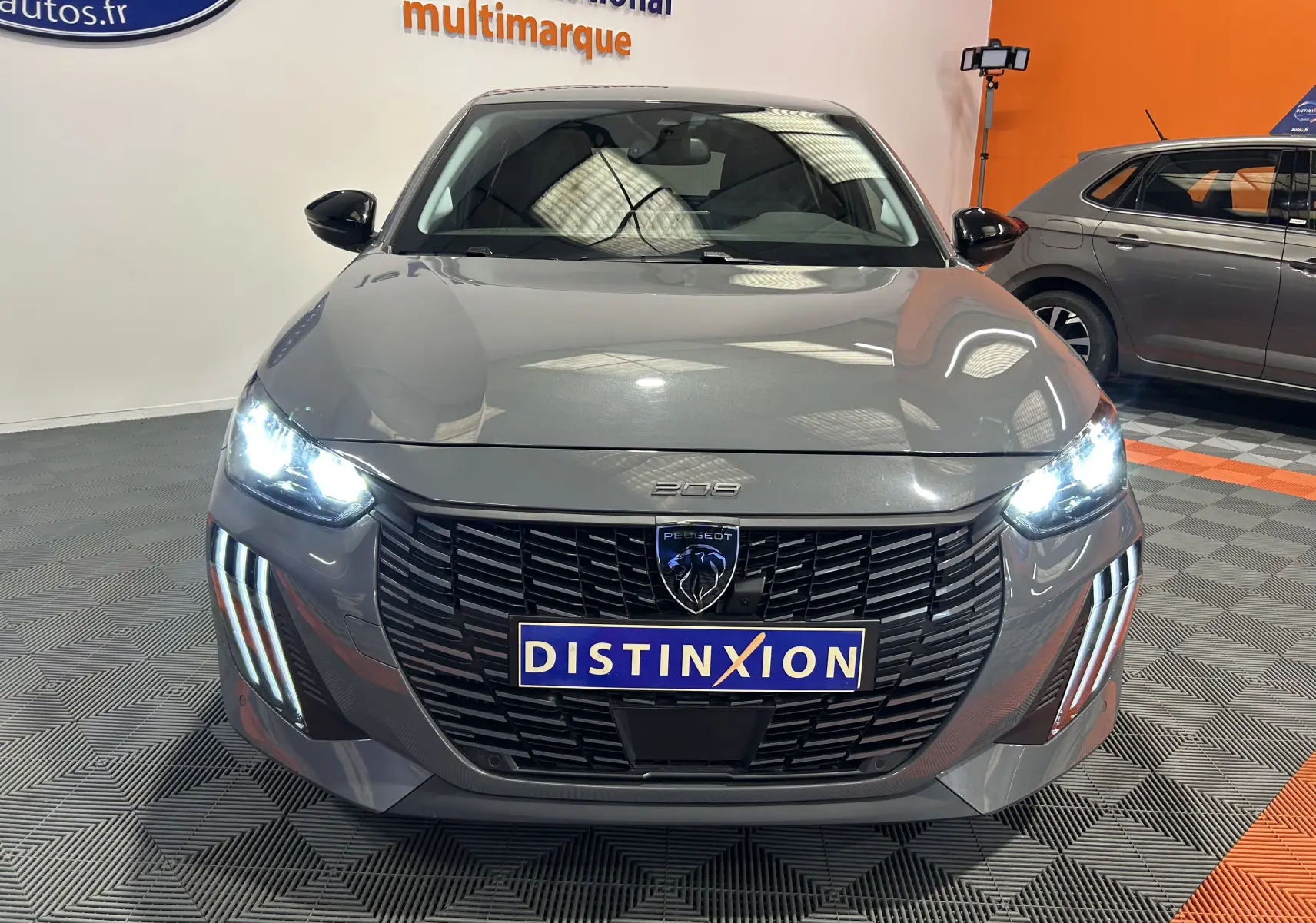 Vue frontale d'une Peugeot 208 gris Selenium avec phares allumés et calandre noire distinctive en showroom.