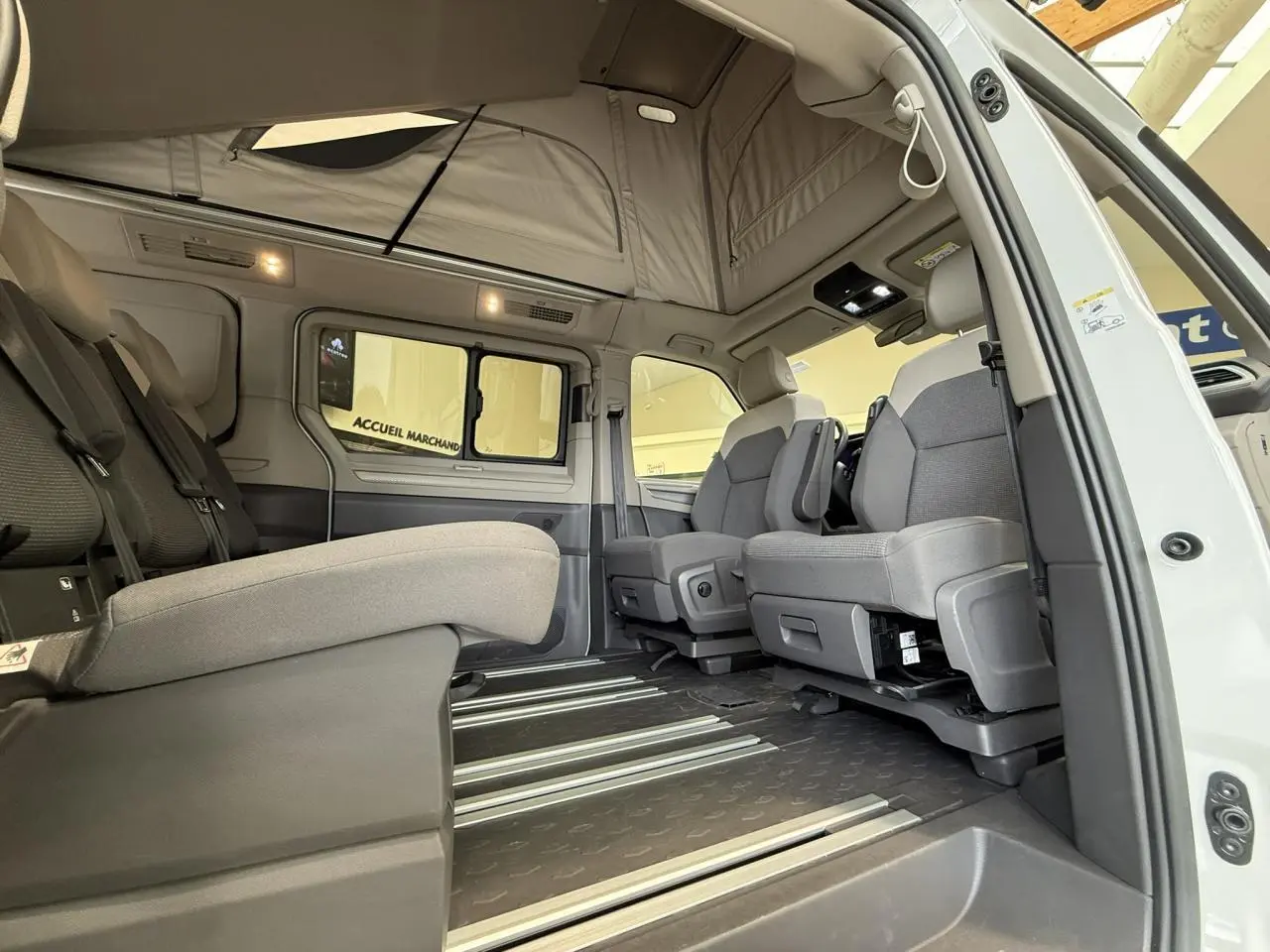 Vue intérieure côté droit du Volkswagen California T7 blanc Candy avec sièges gris et toit relevable ouvert.