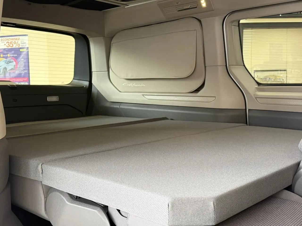 Vue intérieure arrière du Volkswagen California T7 blanc Candy montrant la couchette dépliée et la fenêtre latérale.