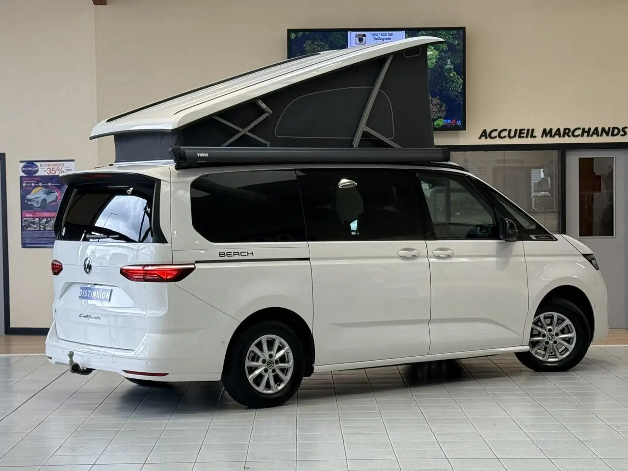 Vue latérale droite d’un Volkswagen California T7 blanc Candy avec toit relevable noir ouvert en intérieur showroom.