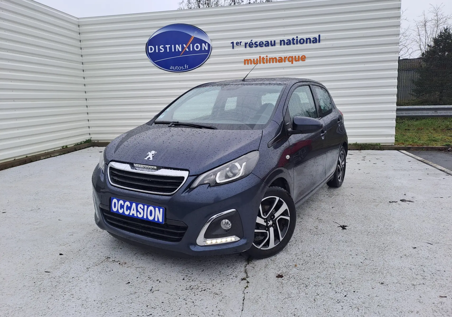 Peugeot 108 bleu vue 3/4 avant droit, avec jantes bi-ton et feux LED allumés sur parking extérieur humide.