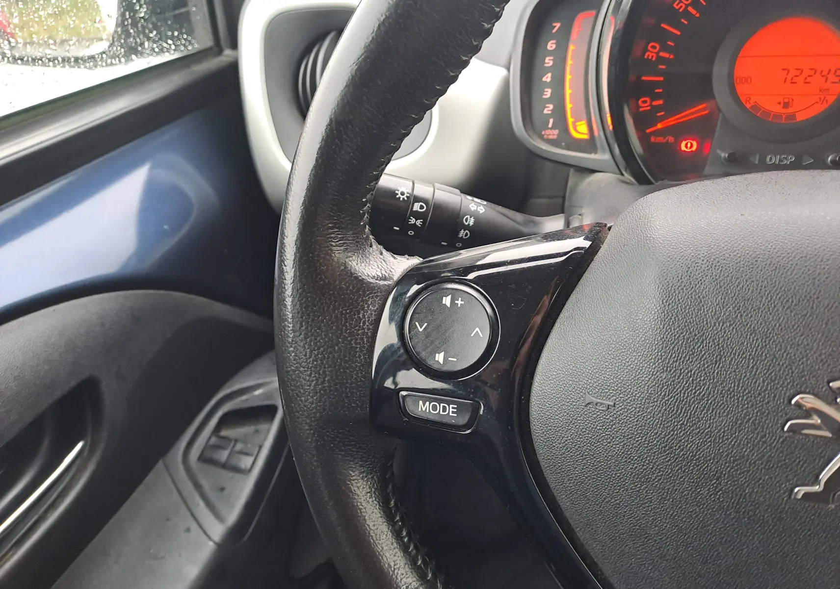 Vue rapprochée du volant côté gauche d'une Peugeot 108 bleue avec commandes audio intégrées et tableau de bord allumé.
