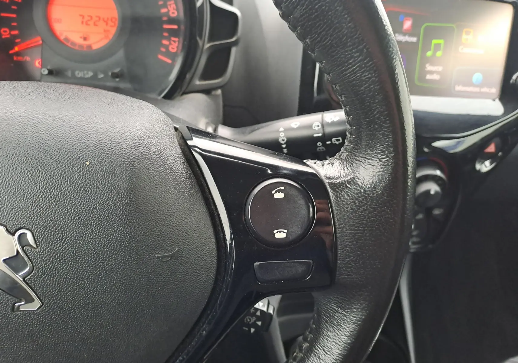 Gros plan sur le volant noir de la Peugeot 108 bleu, avec commandes téléphoniques et tableau de bord éclairé en rouge.