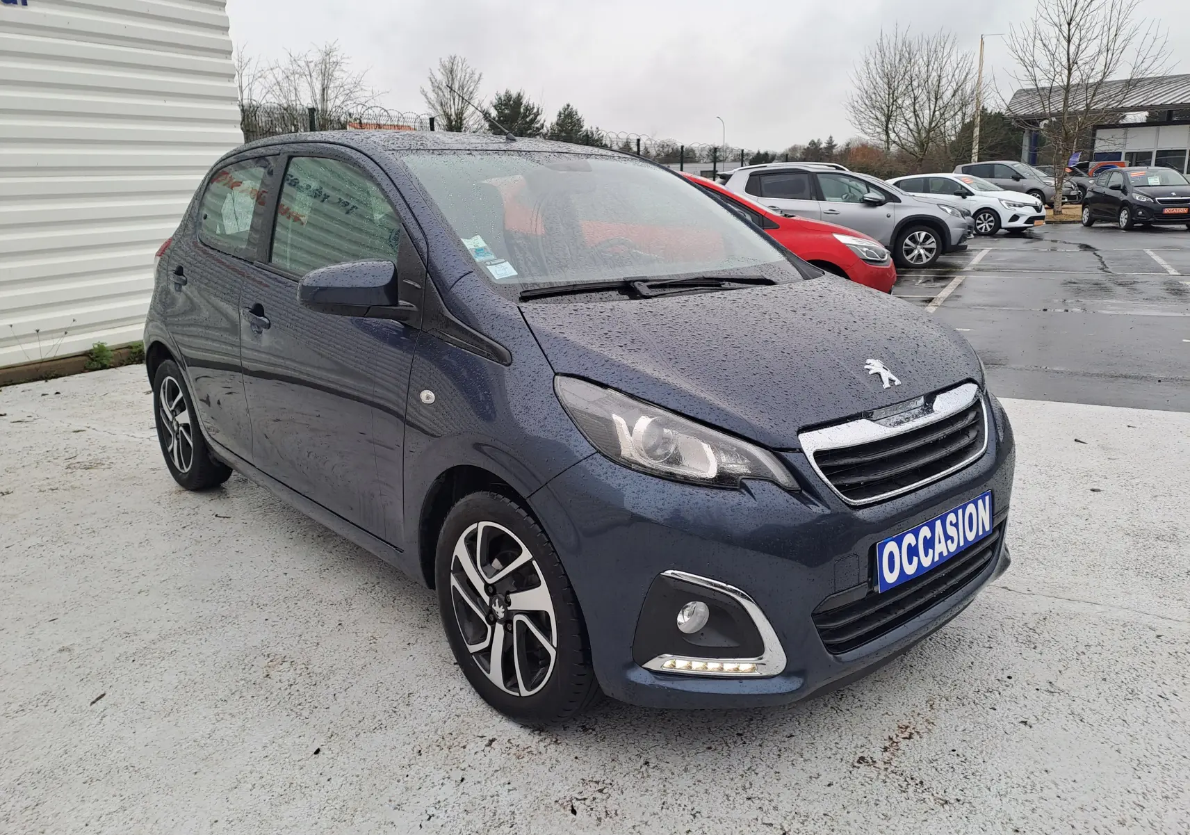 Peugeot 108 bleu vue 3/4 avant gauche sous la pluie avec jantes alliage bicolores et feux LED diurnes allumés.
