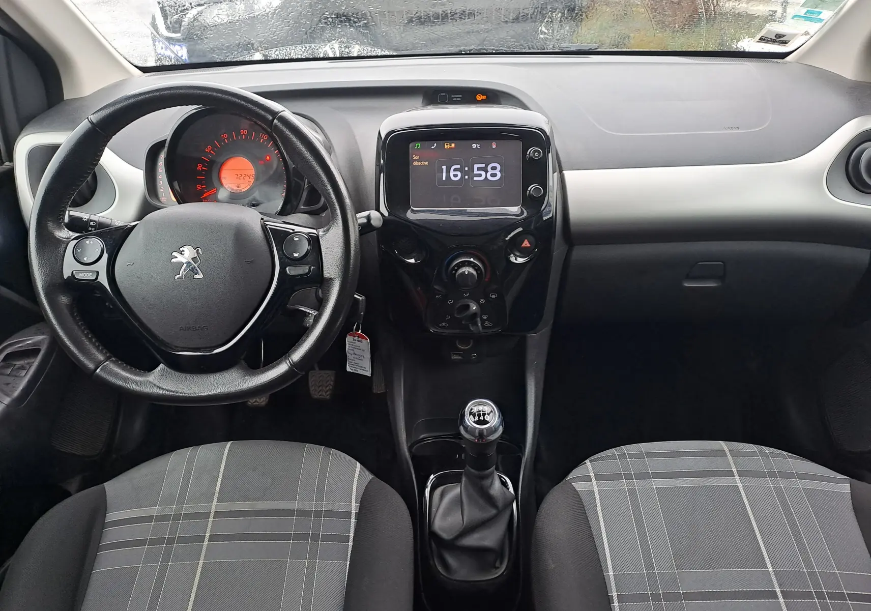 Vue intérieure avant du tableau de bord et volant noir de la Peugeot 108 1.2 82 Allure, avec levier de vitesse manuel et sièges à motif gris.