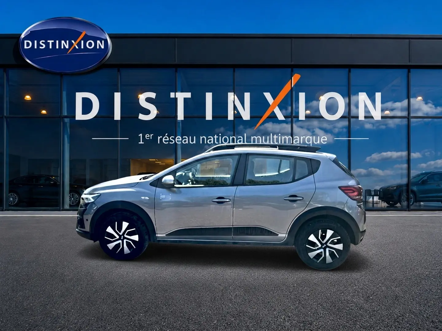 Profil droit d'une Dacia Sandero Stepway gris schiste avec barres de toit, garée devant une vitrine Distinxion.