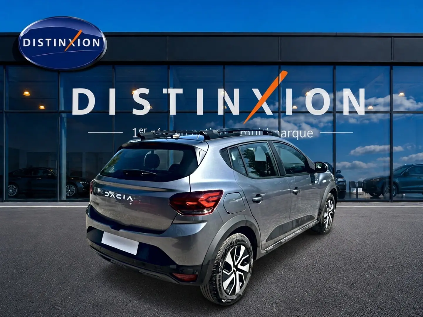 Vue 3/4 arrière droite d'une Dacia Sandero Stepway gris schiste avec barres de toit et jantes alliage devant une vitrine Distinxion.