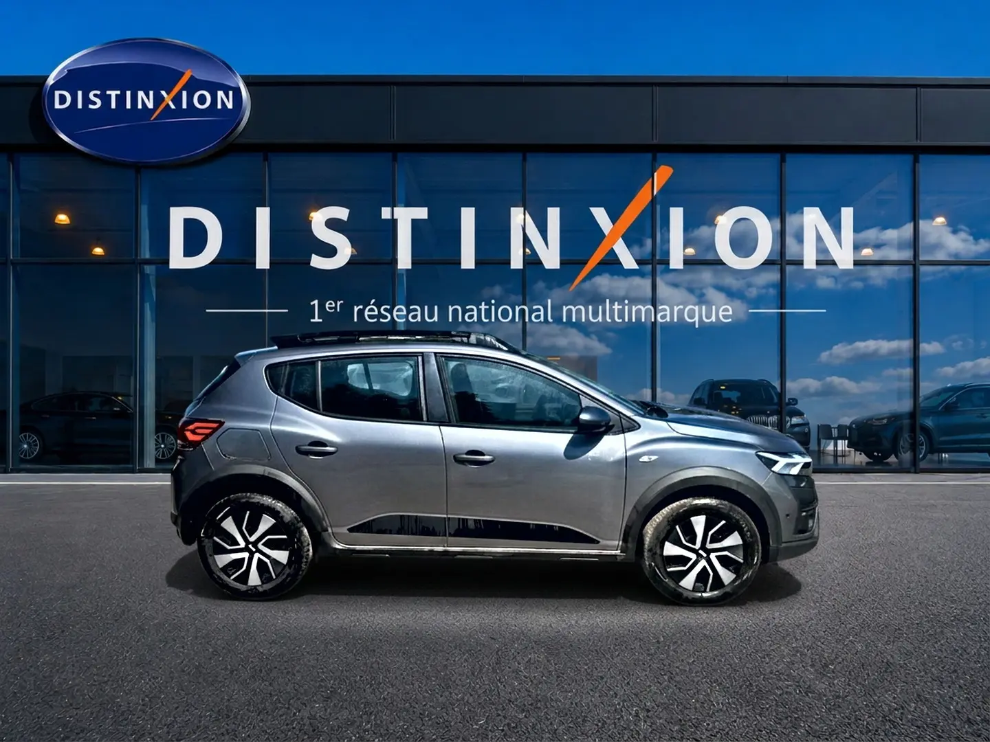 Profil côté gauche d'une Dacia Sandero Stepway gris schiste avec barres de toit et jantes distinctives, devant une vitrine Distinxion.