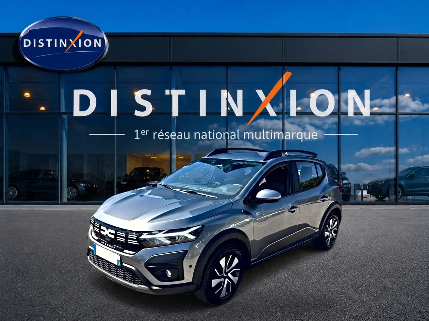 Vue 3/4 avant droit d'une Dacia Sandero Stepway gris schiste avec barres de toit et jantes distinctives, devant une vitrine Distinxion.