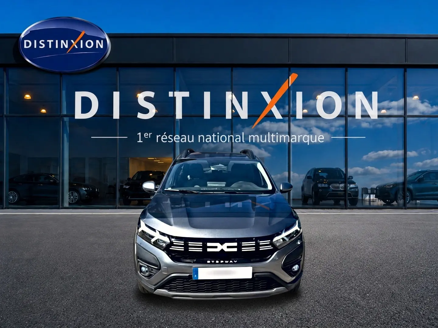 Vue frontale d'une Dacia Sandero Stepway gris schiste avec calandre noire et barres de toit devant un showroom Distinxion.
