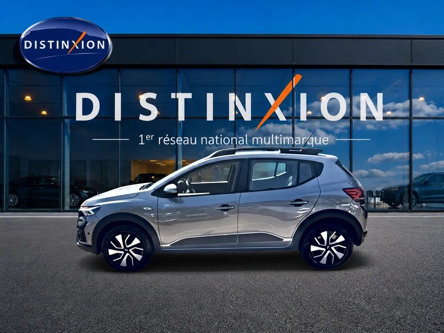 Profil droit de la Dacia Sandero Stepway gris schiste 2025 avec barres de toit et jantes distinctives, devant une vitrine Distinxion.