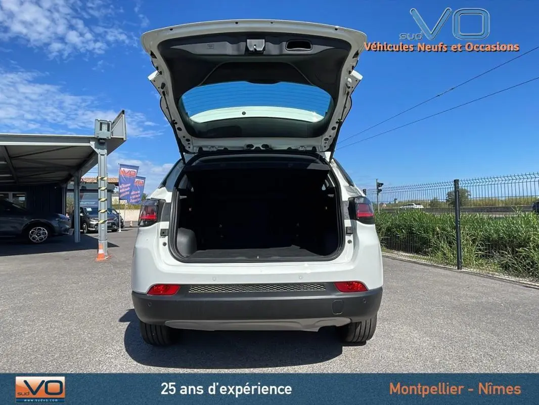Vue arrière d'un Jeep Compass blanc 2024 avec coffre ouvert, sur parking extérieur ensoleillé.