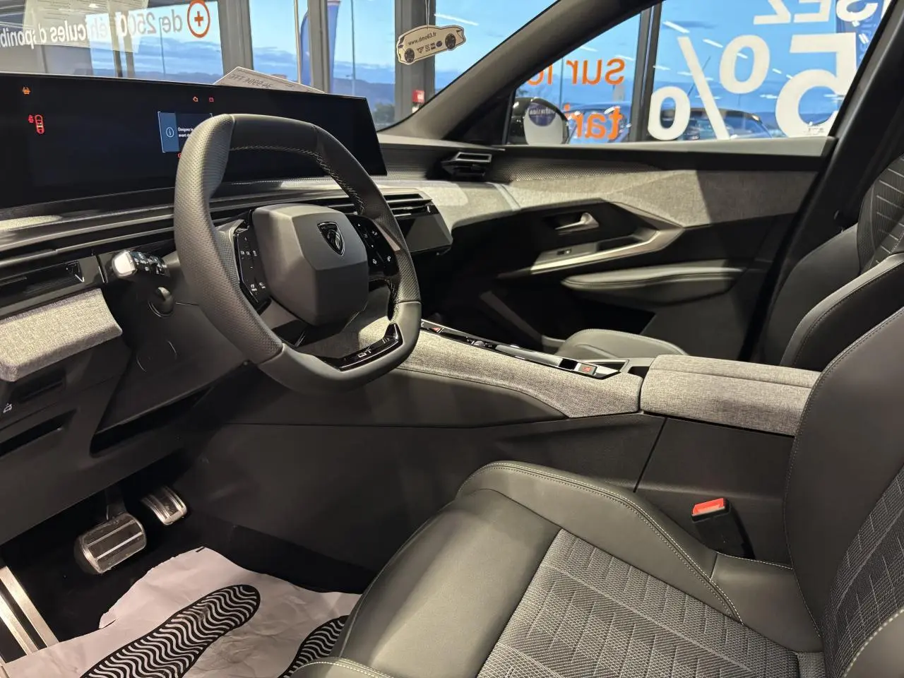 Vue intérieure côté conducteur du Peugeot 3008 GT 2025, avec volant compact, sellerie tissu gris et décor tissu chiné.