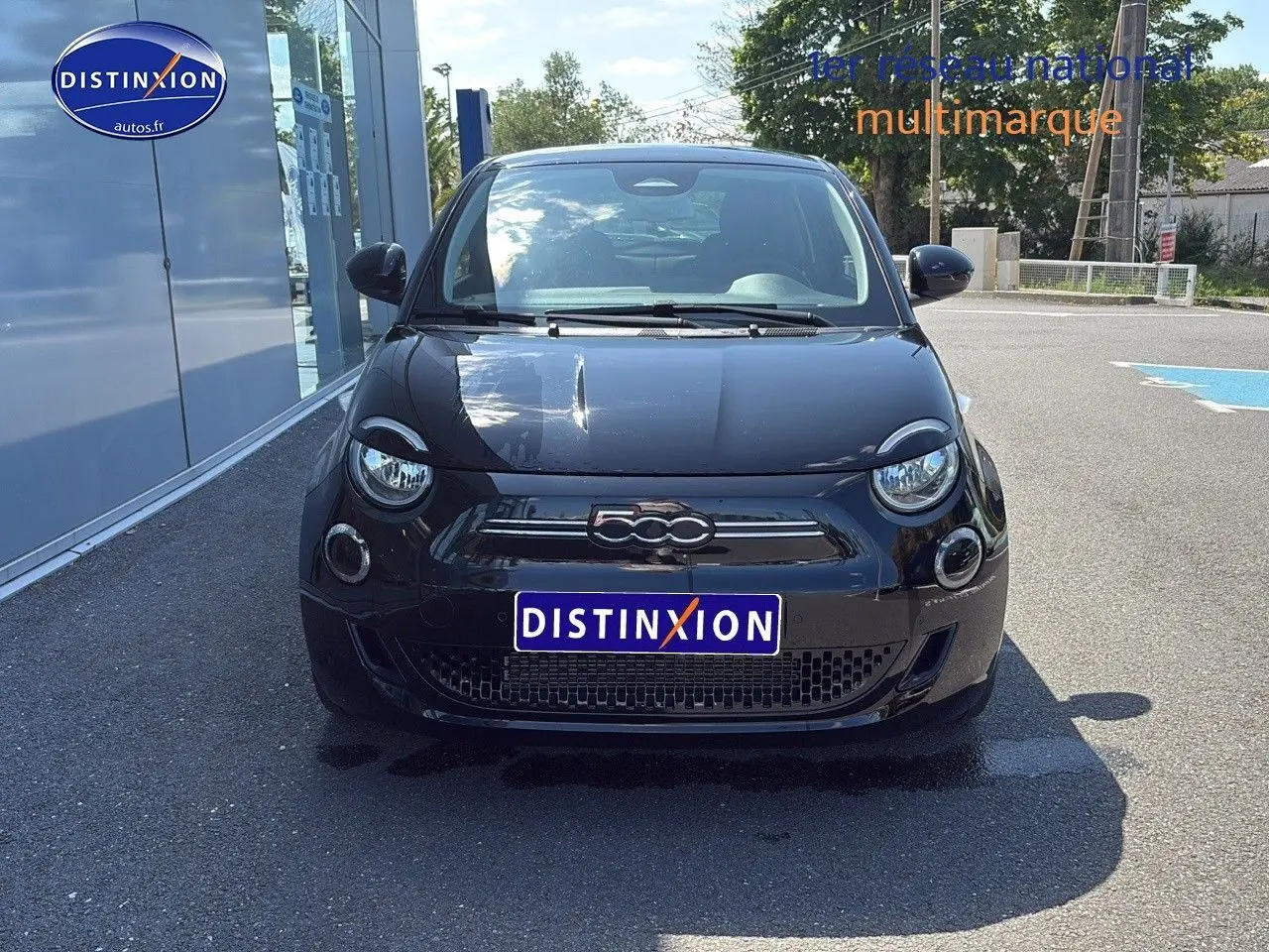 Vue avant d'une FIAT 500 électrique noire avec logo 500 chromé et phares ronds LED, stationnée devant un bâtiment.