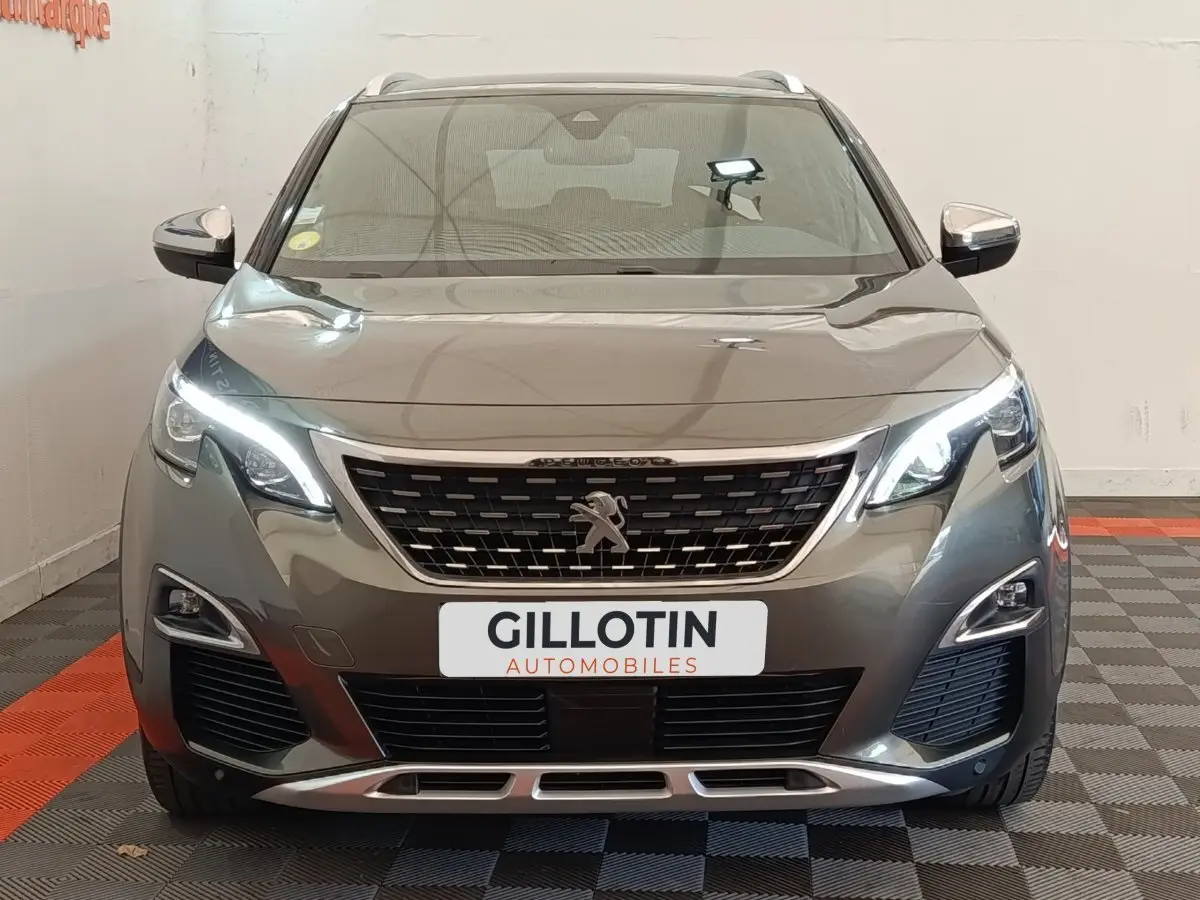 Vue frontale d'un Peugeot 5008 gris clair avec calandre distinctive et feux LED allumés en intérieur showroom.