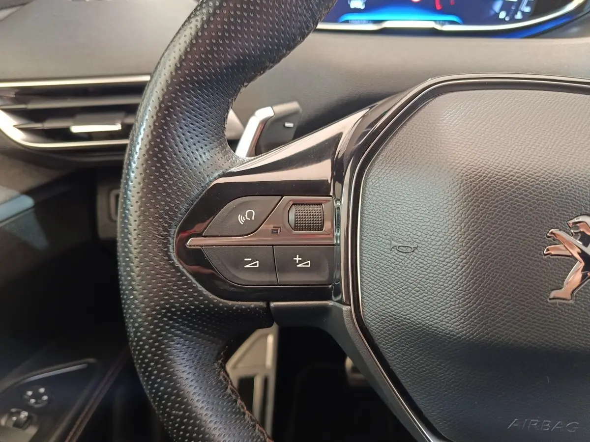 Gros plan sur le volant cuir noir du Peugeot 5008 gris clair, avec commandes audio et logo lion visible.
