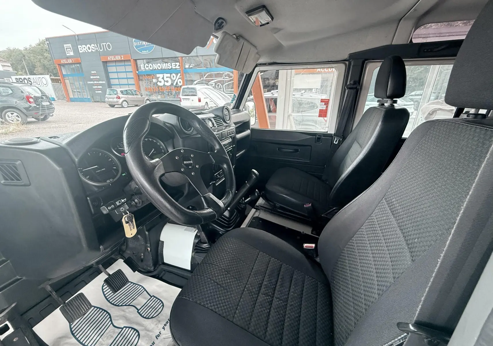 Vue intérieure côté conducteur du Land Rover Defender 2.4 TD4 2007 avec sièges tissu gris-noir et volant sport noir.