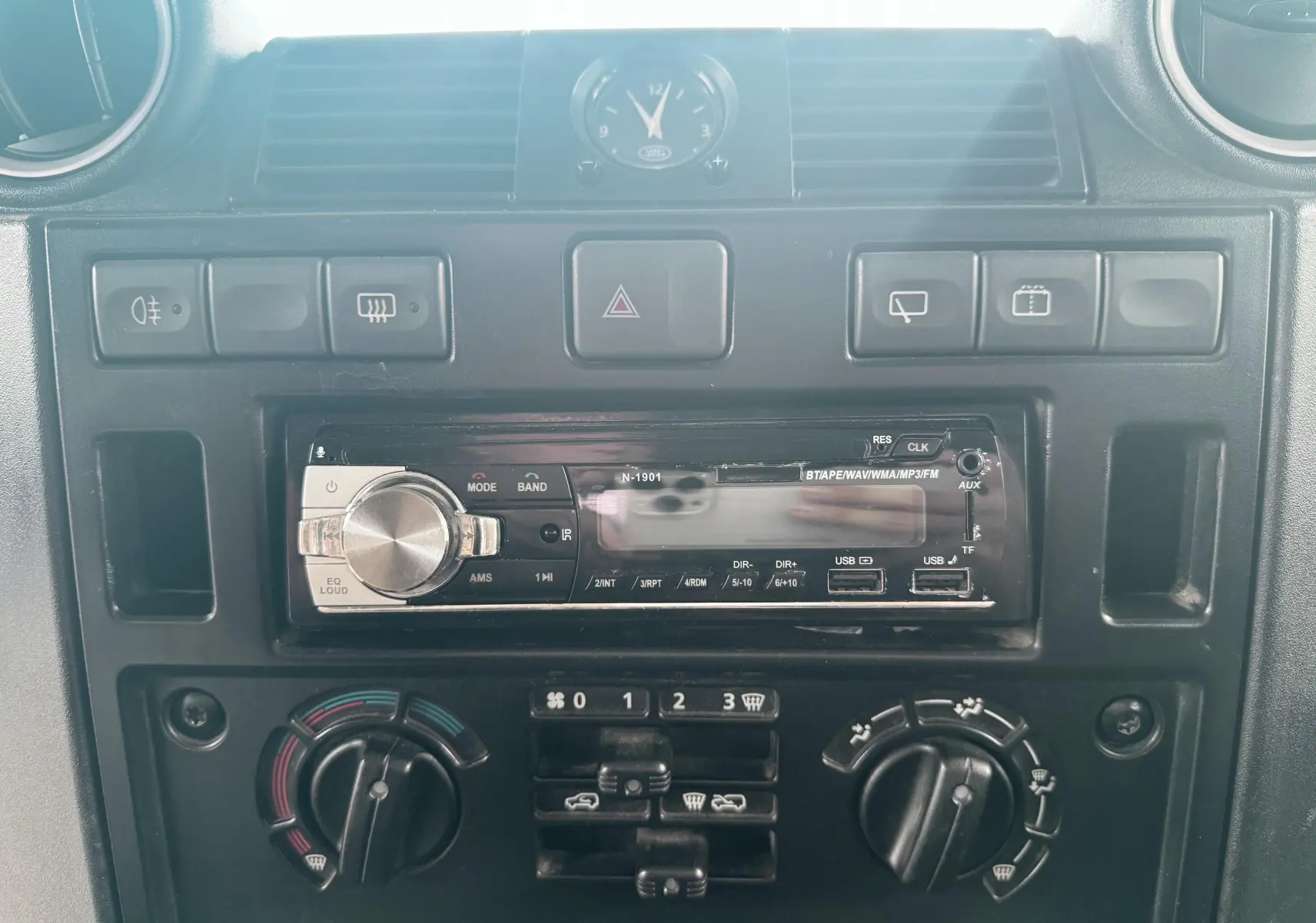 Gros plan sur la console centrale du Land Rover Defender blanc 2007, montrant la radio avec ports USB et commandes de climatisation.
