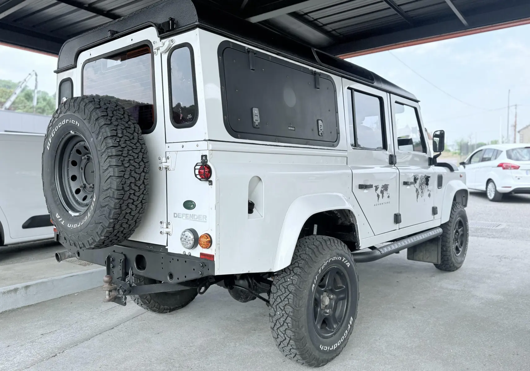 Land Rover Defender blanc vu 3/4 arrière droit, avec toit noir, roue de secours et stickers "Adventure" sur porte.