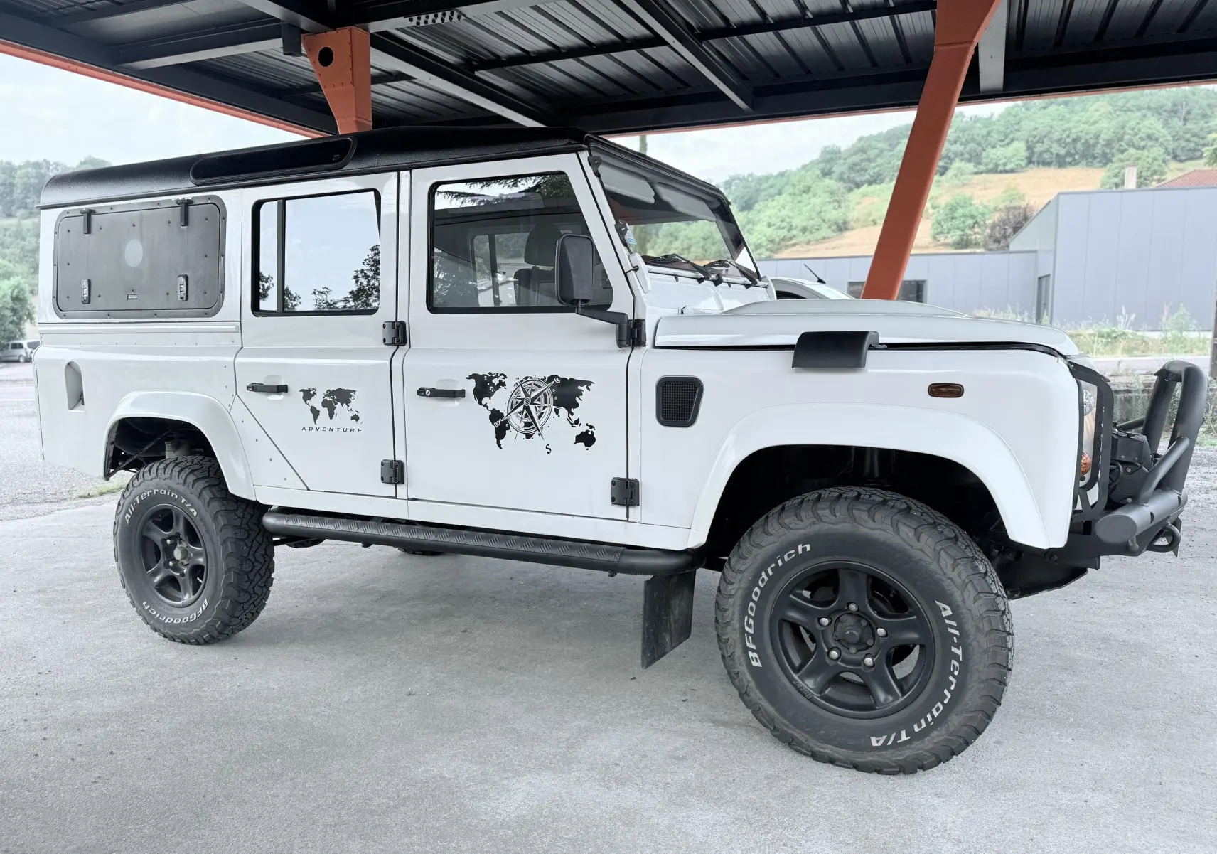Vue de profil droit d'un Land Rover Defender blanc 5 portes avec stickers "Adventure" et roues tout-terrain noires.