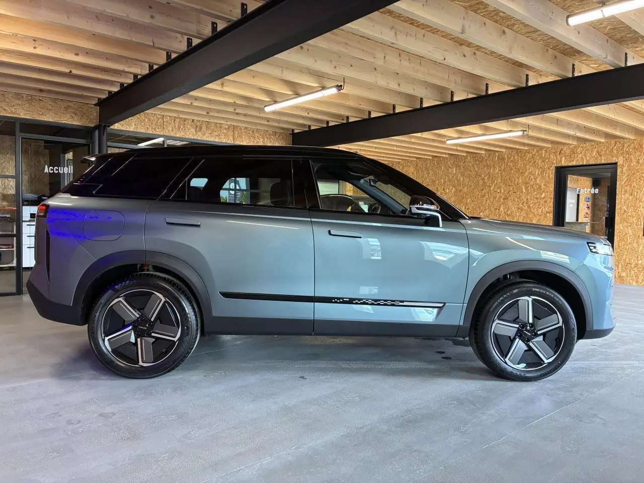 SUV JAECOO J7 2026 gris olive avec toit noir, vu de profil côté gauche dans un garage lumineux.
